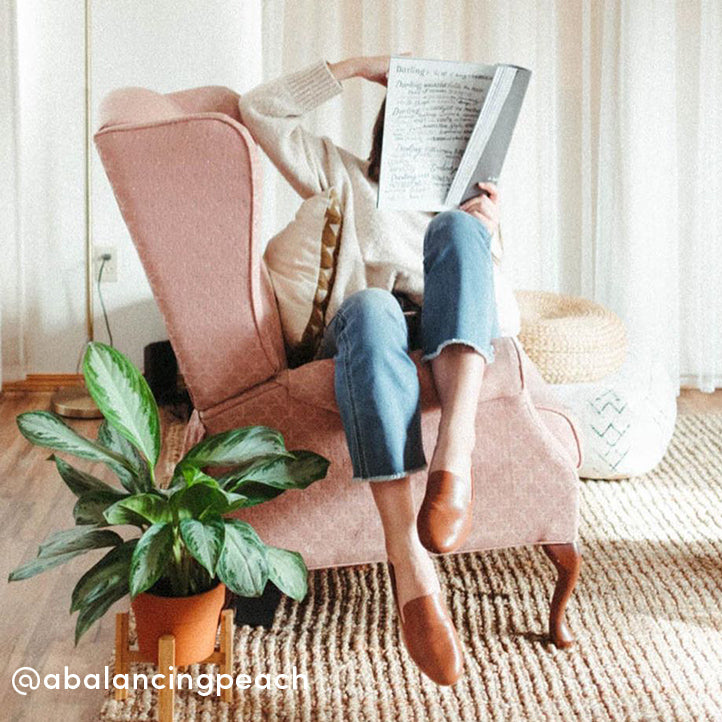 Woman reading a newspaper in a pink chair wearing Birdies’ Starling flats in cognac leather